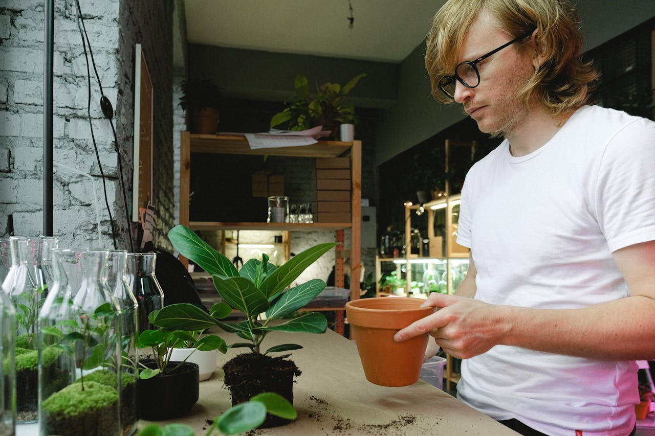 A man indoors potting a plant, surrounded by gardening tools and greenery.